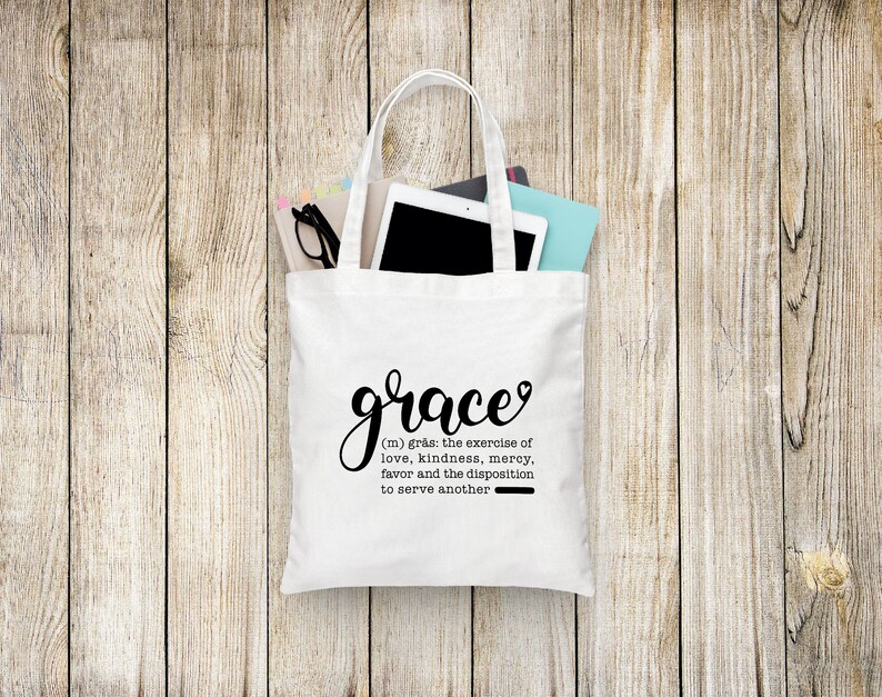May include: White tote bag with black handles and the word "grace" written in cursive with a heart above the e. The definition of grace is written below in black text: "(m) gr&aring;s: the exercise of love, kindness, mercy, favor and the disposition to serve another."
