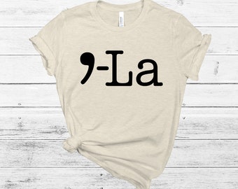 以下が含まれることがあります： ベージュのTシャツに、黒いグラフィックで「9-La」と書いてあります。