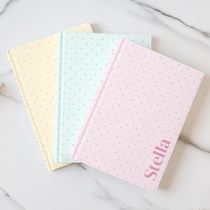 May include: Three hardcover journals with a polka dot pattern in pastel yellow, light blue, and soft pink. The pink journal features the name Stella printed in a dark pink serif font on the front cover. Each notebook has a spine.