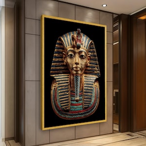 May include: A framed artwork depicting the golden burial mask of Tutankhamun. The mask is set against a black background, with gold, blue, and red accents. The frame is gold. The artwork is displayed on a wall.