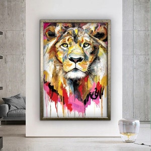 May include: A colorful painting of a lion's face with a silver frame. The lion's fur is painted in shades of yellow, orange, and brown, with splashes of pink, red, and blue paint around the face. The painting has a white background.