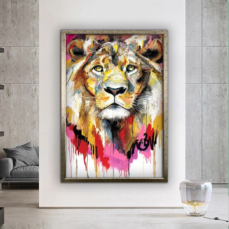 May include: A colorful painting of a lion's face with a silver frame. The lion's fur is painted in shades of yellow, orange, and brown, with splashes of pink, red, and blue paint around the face. The painting has a white background.