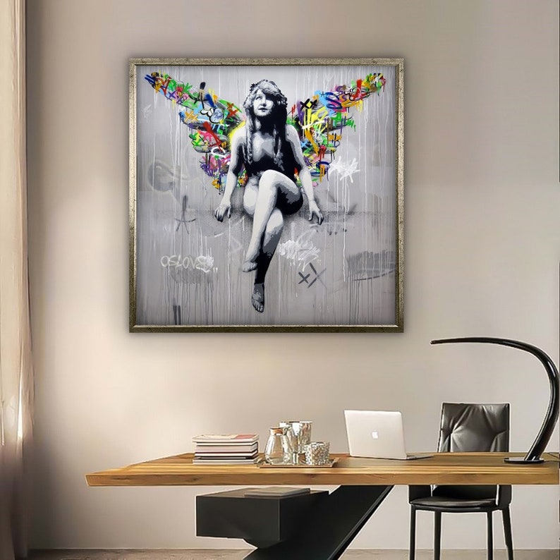 May include: A framed print of a woman with colorful wings sitting on a gray background. The wings are made of graffiti-style lettering and the woman is wearing a white dress. The print is framed in a silver frame.