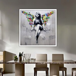 May include: A framed print of a woman with wings, sitting on a gray background with colorful graffiti. The woman is wearing a white dress and has long, dark hair. The graffiti includes the word "of love".