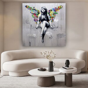 May include: A colorful abstract painting of an angel with graffiti-style wings. The angel is sitting with its legs crossed and has a halo. The background is a white canvas with black and gray paint splatters.
