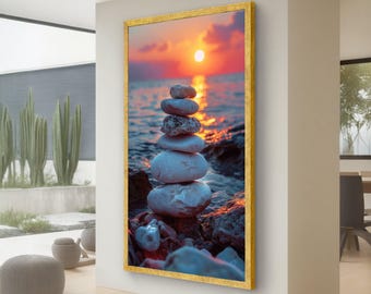 Puede incluir: Impresión enmarcada que presenta una pila de piedras lisas y claras equilibradas en una costa rocosa. El fondo muestra una vibrante puesta de sol sobre el océano, con el sol reflejado en el agua. El marco es dorado.
