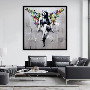 May include: A black and white image of a woman with wings made of colorful graffiti. The woman is sitting on a ledge with her legs crossed. The background is a gray wall with drips of paint.