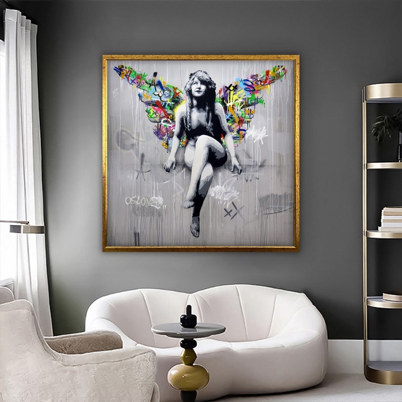 May include: A framed canvas print of a woman with wings made of colorful graffiti. The woman is sitting with her legs crossed and her arms resting on her knees. The background is a light gray with a white and gray paint splatter effect. The frame is gold.
