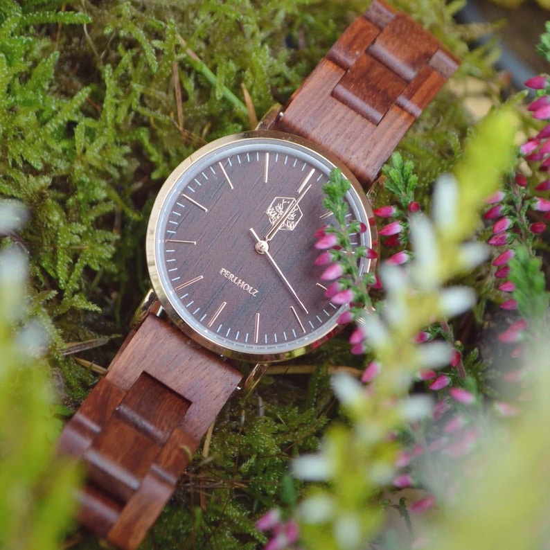 May include: A wooden wristwatch with a dark brown face and band. The watch has a gold-colored bezel and hands. The watch face has the text "PERLHOLZ" and is surrounded by a gold-colored ring. The watch is surrounded by green moss and pink flowers.