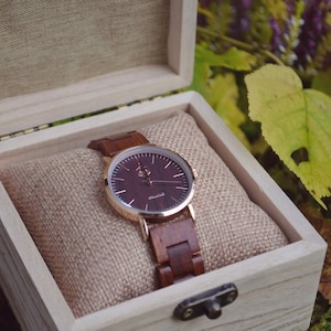 May include: A wooden wristwatch with a dark brown face and matching wooden band, presented in a light-colored wooden box. The watch has gold-colored accents and is displayed on a tan fabric cushion.