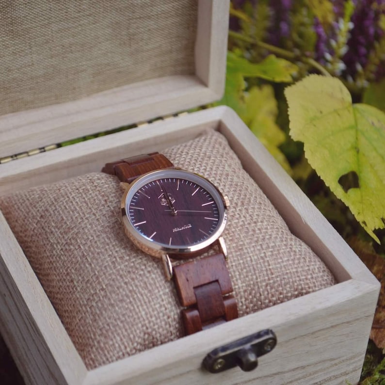 May include: A wooden wristwatch with a dark brown face and matching wooden band, presented in a light-colored wooden box. The watch has gold-colored accents and is displayed on a tan fabric cushion.