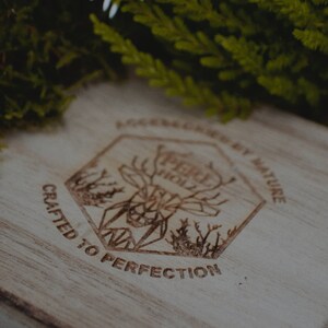 May include: Close-up of a wooden surface with a laser-etched design. The design features a geometric deer head within a hexagonal frame, surrounded by text that reads "PERI HOLZ" and "CRAFTED TO PERFECTION". Green foliage is visible in the background.