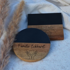 Puede incluir: Dos posavasos de madera con tapas de resina epoxi negra. Un posavasos es redondo y presenta el texto "Familie Eckhardt" y un diseño de dos manos sosteniendo un corazón. El otro posavasos es cuadrado y presenta el texto "die Kaletsch" y un icono de casa.