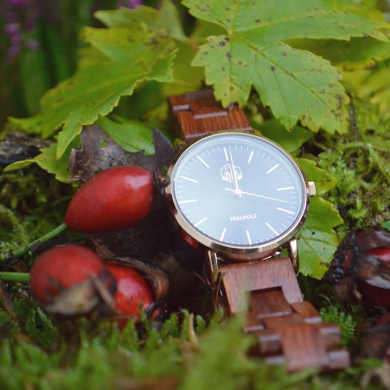 May include: A wristwatch with a dark gray face and gold-colored bezel. The watch has a wooden band and is surrounded by green foliage and red berries. The watch face has the text "PERLHOLZ".