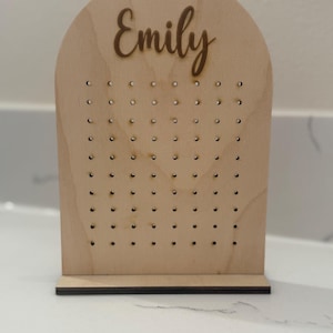 May include: A wooden earring holder with a rounded top and a base. The holder has a name, "Emily", engraved on the top and a grid of holes for holding earrings.
