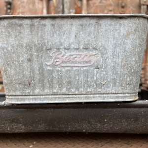 May include: A rectangular, galvanised metal tub with a slightly tapered design. The word "Beau" is embossed in a cursive font on the front. The tub has a weathered appearance, suggesting age and use. The tub is sitting on a black surface.