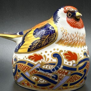 May include: A decorative ceramic bird figurine. The bird is predominantly white, with intricate detailing in red, blue, gold, and orange. The base features a pattern in blue, gold, and red. The bird's head is red and white.