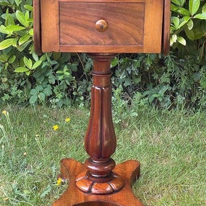 Op de afbeelding: Een donkerbruine antieke bijzettafel met een lade en een ronde knop. De tafel heeft een voetstuk en een vierpotige basis. De tafel is gemaakt van hout en staat op een grasachtig oppervlak.