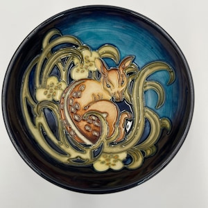 May include: A ceramic bowl with a detailed design. The bowl features a sleeping fawn in shades of brown and cream, surrounded by stylized floral and plant motifs in green and cream, set against a blue background. The rim is dark blue.