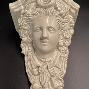 May include: A light grey ceramic wall sconce depicting a classical female face with detailed hair, a crescent moon, and floral accents. The sculpture has a textured finish and is designed for wall mounting.
