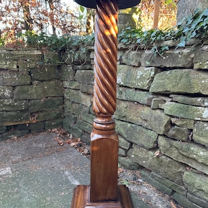 May include: A dark brown wooden pedestal stand with a round top and a twisted column design. The base is square and the stand is outdoors, in front of a stone wall.