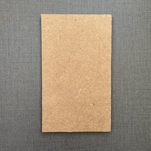 May include: A rectangular, light brown particle board panel with a rough, textured surface. The panel is set against a dark grey, textured background.