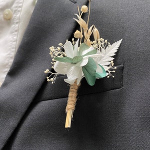 Peut inclure: Un bouton de fleur composé de fleurs en papier blanches et vertes, de blé séché et de délicats accents blancs. Les tiges sont enveloppées de ficelle et de paille, fixées à une veste de costume noire et une chemise blanche.