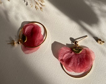 May include: Gold-coloured hoop earrings featuring a petal design in a soft, dusty rose hue. The earrings are set against a neutral background, with natural light creating shadows.