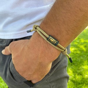 Pode incluir: Um bracelete trançado bege e verde com um detalhe em metal preto e uma inscrição dourada. O bracelete é usado no pulso de uma pessoa.