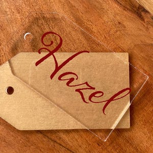 May include: A clear acrylic tag with the name "Hazel" in red script, layered over a brown paper tag. Both tags have a hole for hanging. The tags are on a wooden surface.