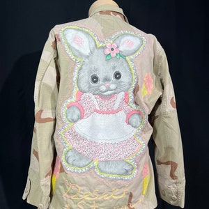 May include: A camouflage jacket with a pink and grey bunny embroidered on the back. The bunny is wearing a pink dress with white trim and a pink flower in its hair. The text "Desert Storm" is embroidered below the bunny.