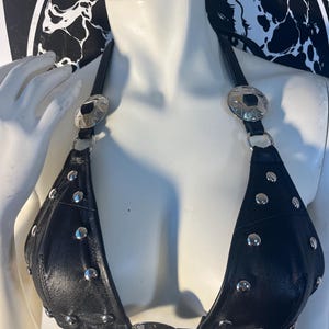 May include: Black leather bikini top with silver-tone metal studs and decorative accents. The top features a halter neck design with large silver-tone metal embellishments at the straps. The triangle cups are adorned with multiple silver studs.