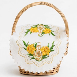 May include: A wicker Easter basket with a handle, featuring a white scalloped cloth. The cloth is embroidered with yellow daffodils, green leaves, and decorated Easter eggs. The basket is designed for Easter celebrations.