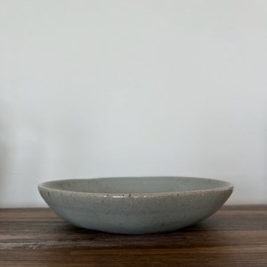 May include: A light grey ceramic bowl with a slightly textured surface. The bowl is round and shallow, with a wide rim.