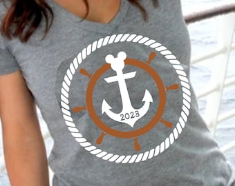 May include: Gray v-neck t-shirt featuring a nautical design with a white rope circle, a brown ship's wheel, and a white anchor with the year 2023. The shirt is a casual style.