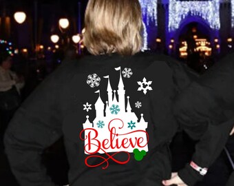 May include: Two people wearing black long-sleeved shirts with a white silhouette of Cinderella's castle and the word "Believe" in red. The castle is decorated with snowflakes and a green Mickey Mouse head.