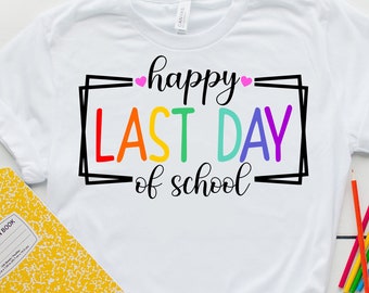 Op de afbeelding: Wit T-shirt met de tekst "happy last day of school" in regenboogkleuren en roze hartjes. Een geel schrift en kleurpotloden liggen op een wit houten oppervlak.