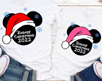May include: Two white t-shirts with a black Mickey Mouse head design. One shirt has a red Santa hat and the other has a pink Santa hat. Both shirts have the text "Disney Christmas 2022".