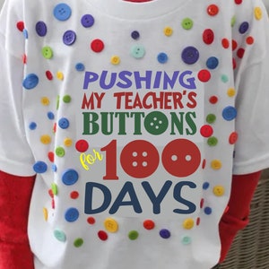 Puede incluir: Camiseta blanca decorada con botones coloridos y la frase "PUSHING MY TEACHER'S BUTTONS for 100 DAYS" en varios colores. La camiseta tiene una camiseta roja de manga larga debajo.