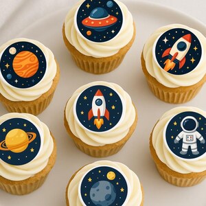 May include: Seven cupcakes with white frosting and space-themed decorations. Each cupcake has a circular image of planets, rockets, an astronaut, and a UFO. The cupcakes are arranged on a white plate, perfect for a space-themed party or event.