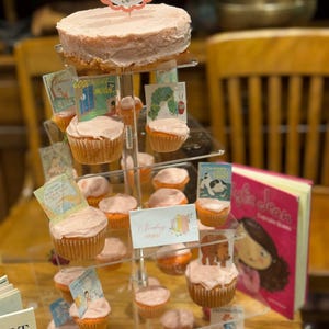 K&ouml;nnte beinhalten: Mehrst&ouml;ckiger Dessertst&auml;nder mit einem gro&szlig;en Kuchen und Cupcakes, alle mit rosa Zuckerguss bedeckt. Miniatur-Buchcover schm&uuml;cken jeden Cupcake. Ein Schild liest "Erdbeer-Cupcakes". Ein rosa Buch ist ebenfalls sichtbar.