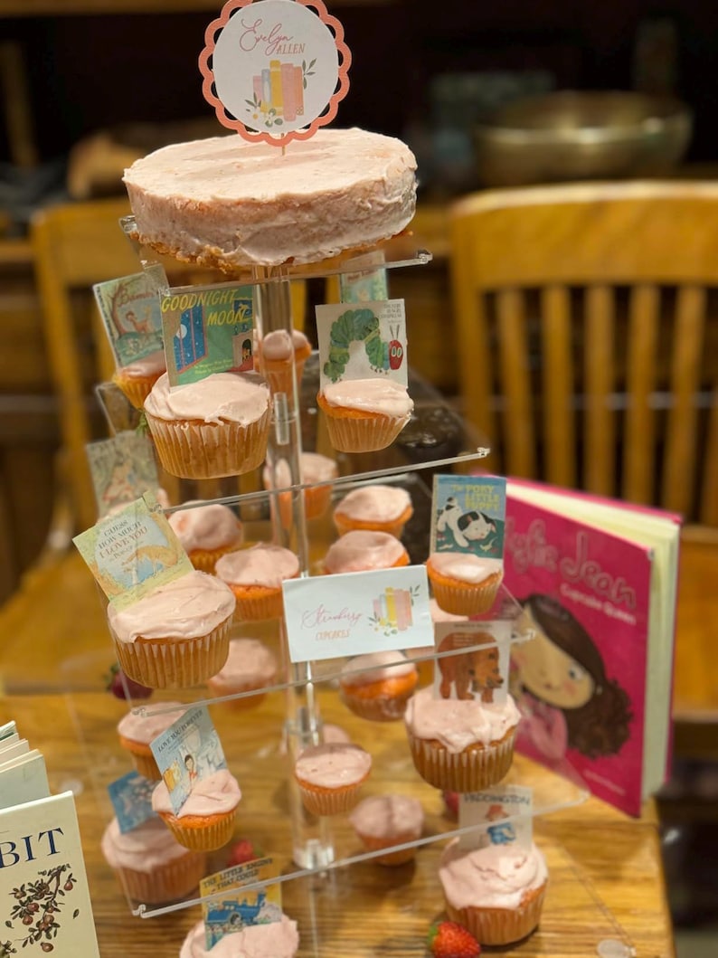 K&ouml;nnte beinhalten: Mehrst&ouml;ckiger Dessertst&auml;nder mit einem gro&szlig;en Kuchen und Cupcakes, alle mit rosa Zuckerguss bedeckt. Miniatur-Buchcover schm&uuml;cken jeden Cupcake. Ein Schild liest "Erdbeer-Cupcakes". Ein rosa Buch ist ebenfalls sichtbar.