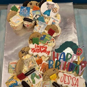 K&ouml;nnte beinhalten: Ein Cupcake-Kuchen, verziert mit Keksen, S&uuml;&szlig;igkeiten und Miniatur-Buch-Toppern. Der Kuchen schreibt "Happy Birthday Jessa" und zeigt eine Raupe und andere Buchfiguren. Der Kuchen ist mit wei&szlig;em Zuckerguss bedeckt.