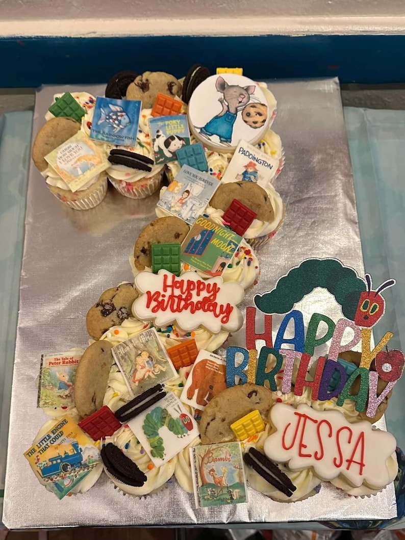 K&ouml;nnte beinhalten: Ein Cupcake-Kuchen, verziert mit Keksen, S&uuml;&szlig;igkeiten und Miniatur-Buch-Toppern. Der Kuchen schreibt "Happy Birthday Jessa" und zeigt eine Raupe und andere Buchfiguren. Der Kuchen ist mit wei&szlig;em Zuckerguss bedeckt.