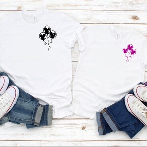 May include: Two white t-shirts with a black and pink Mickey Mouse balloon design. The shirts are laid out on a white wooden surface with blue jeans and white sneakers.