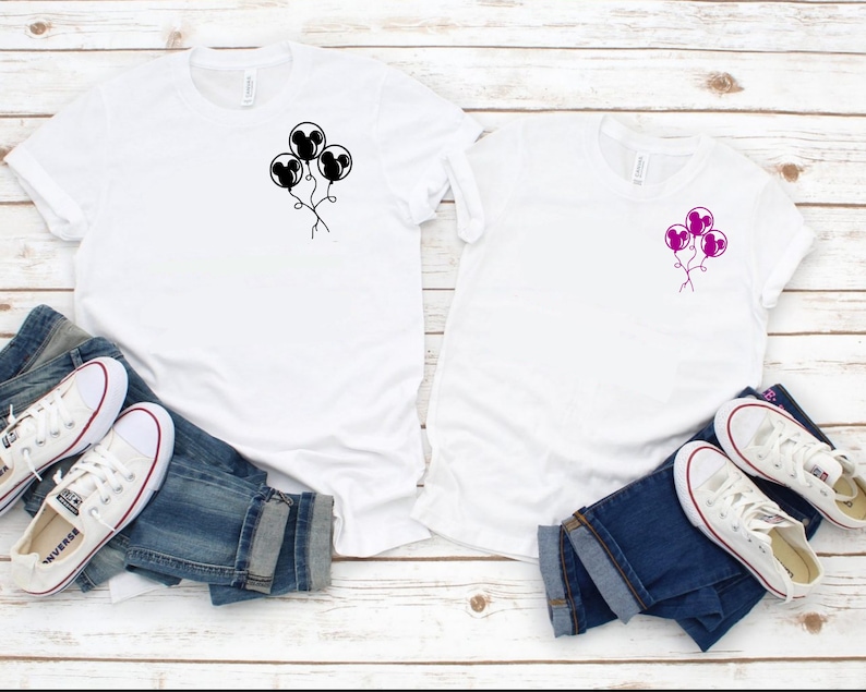 May include: Two white t-shirts with a black and pink Mickey Mouse balloon design. The shirts are laid out on a white wooden surface with blue jeans and white sneakers.