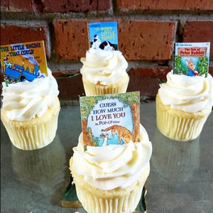 K&ouml;nnte beinhalten: Vier Cupcakes mit wei&szlig;em Zuckerguss und Miniatur-Buch-Toppern. Die B&uuml;cher tragen die Titel "The Little Engine That Could", "The Poky Little Puppy", "The Tale of Peter Rabbit" und "Guess How Much I Love You". Die Cupcakes stehen auf einer reflektierenden Oberfl&auml;che.
