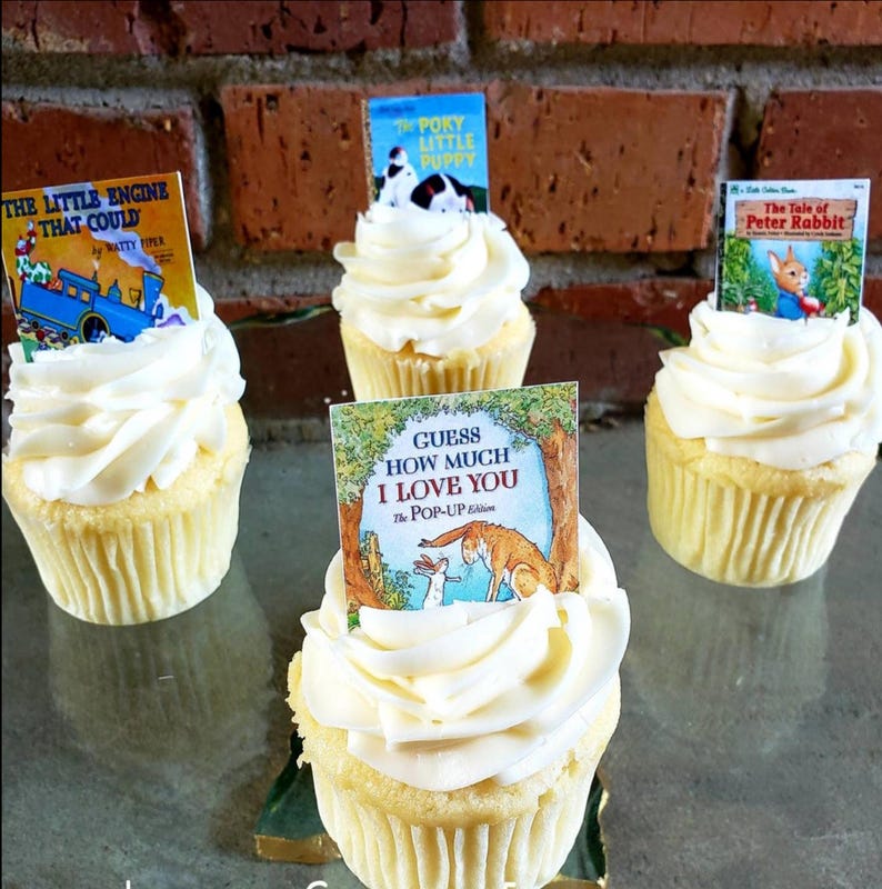 K&ouml;nnte beinhalten: Vier Cupcakes mit wei&szlig;em Zuckerguss und Miniatur-Buch-Toppern. Die B&uuml;cher tragen die Titel "The Little Engine That Could", "The Poky Little Puppy", "The Tale of Peter Rabbit" und "Guess How Much I Love You". Die Cupcakes stehen auf einer reflektierenden Oberfl&auml;che.
