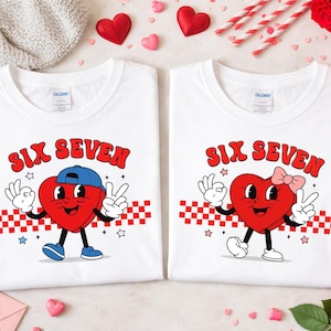 May include: Two white t-shirts with the text "SIX SEVEN" in red. Each shirt features a cartoon heart character with a face, arms, and legs. One heart wears a blue cap and the other a pink bow. A red and white checkered pattern is below the text.
