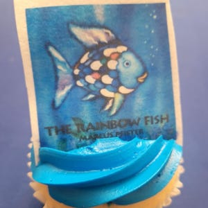 K&ouml;nnte beinhalten: Ein Cupcake mit blauer Glasur und einem rechteckigen essbaren Bild von "The Rainbow Fish" von Marcus Pfister. Das Bild zeigt einen blauen Fisch mit bunten Schuppen vor blauem Hintergrund. Der Cupcake befindet sich in einer wei&szlig;en Papierform.